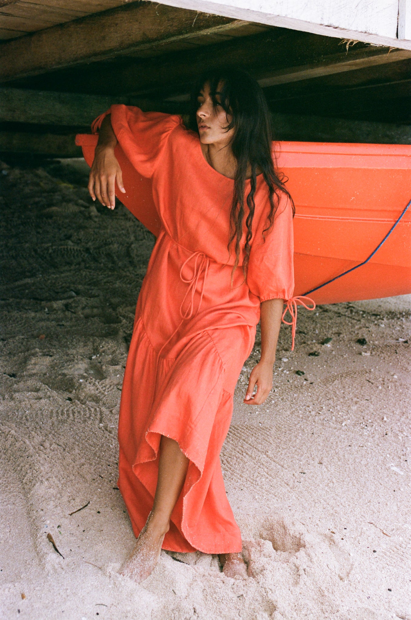 LILLY PILLY woman  wearing Charlotte linen  dress in  Spritz standing under a wooden structure with a red container in the background.
