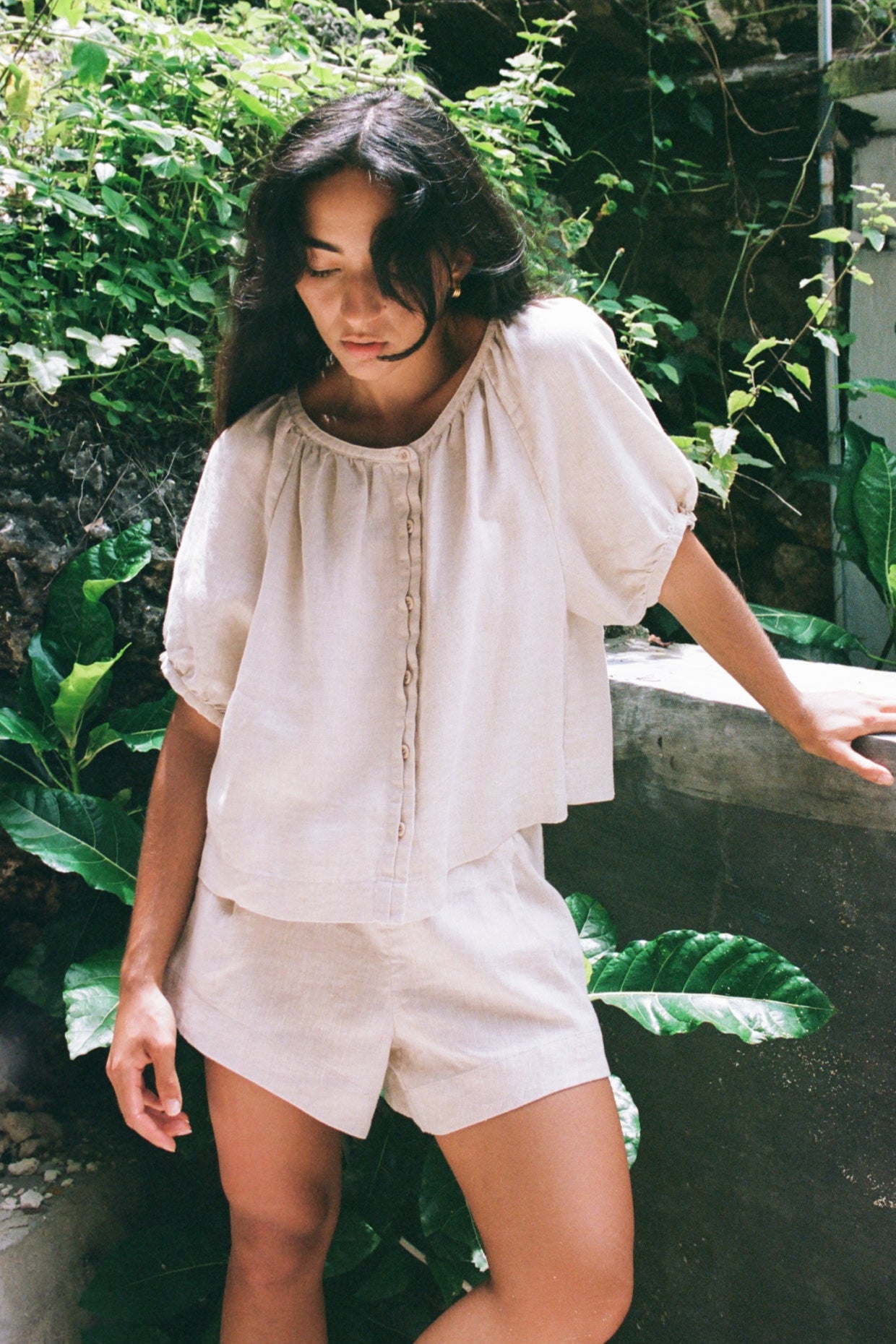 LILLY PILLY woman wearing a oatmeal Pia linen shirt top and Gaia shorts standing in a natural setting with greenery.